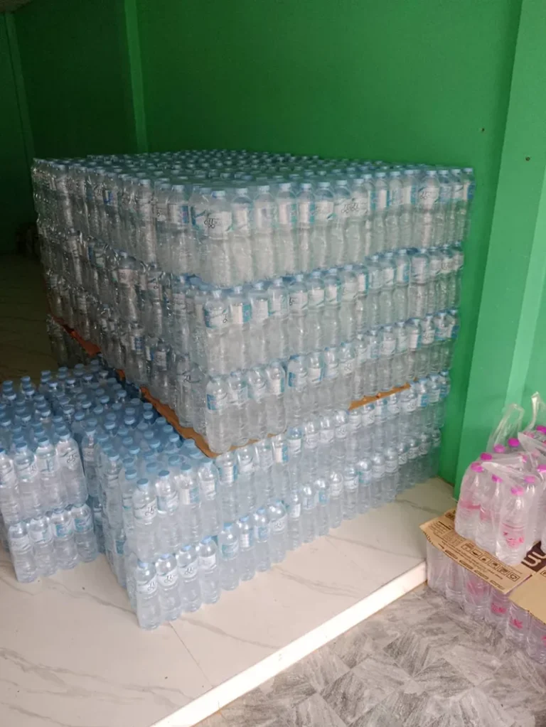 บริการ ส่งน้ำดื่ม มข กังสดาล - หลัง มข. น้ำดื่มราคาส่ง ส่งไวถึงหอพักและร้านอาหาร 11 ขวดน้ำที่บรรจุแล้วเรียงเป็นตั้งๆ และเครื่องจักร ภายในโรงงาน